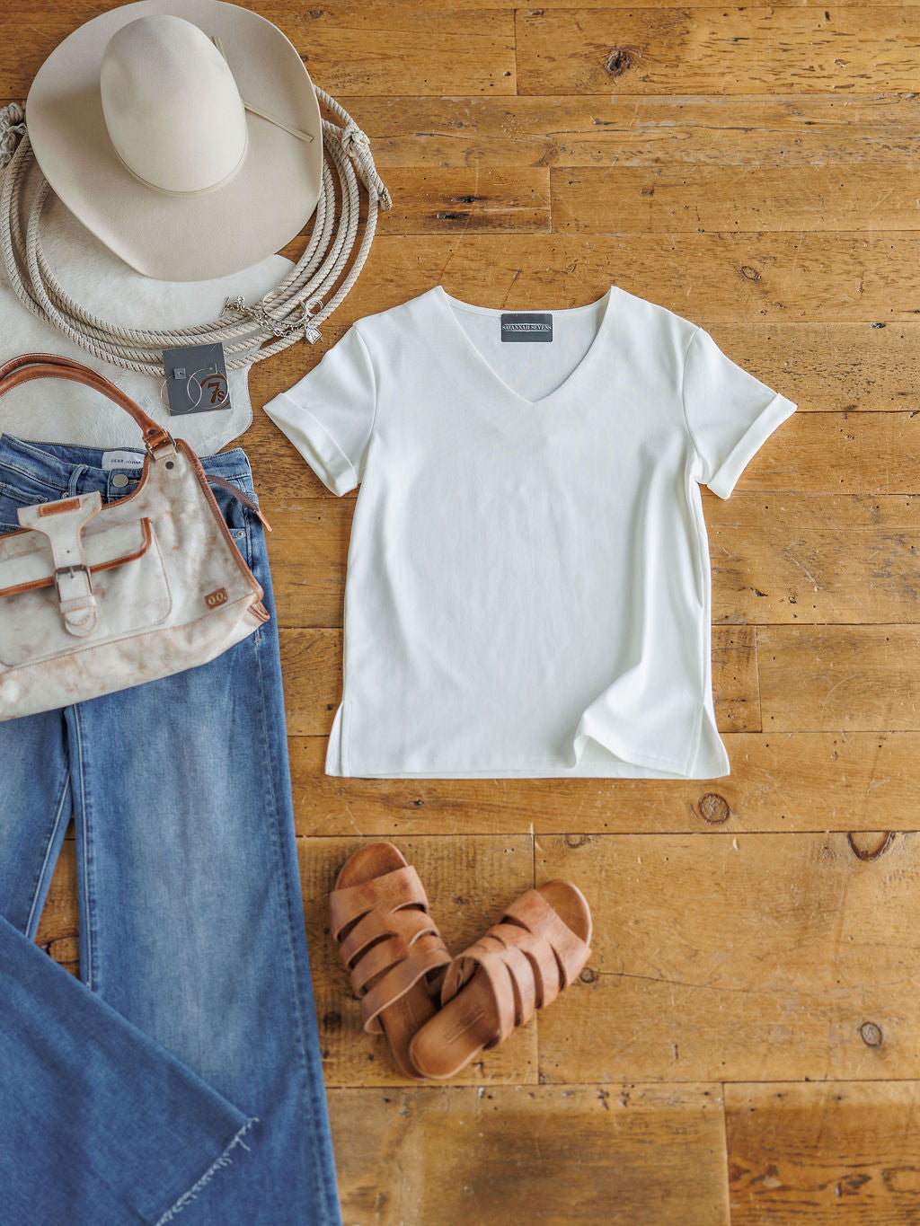 A flat lay on a wooden floor features The Kinston short sleeve top by Savannah Sevens western life{&}style, styled with blue jeans, tan sandals, a cream patterned handbag, white cowboy hat with rope trim, and sunglasses.