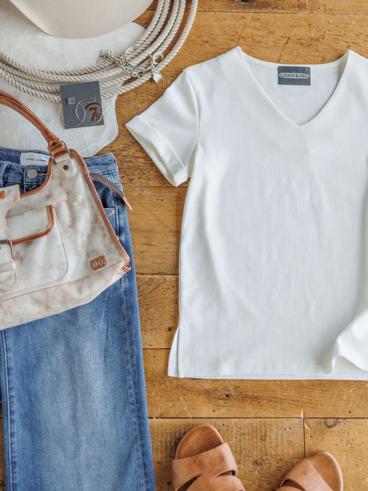 A flat lay featuring The Kinston by Savannah Sevens western life{&}style: an off-white short sleeve top, blue jeans, tan sandals, beige handbag and hat, plus a beaded necklace on a wooden floor.