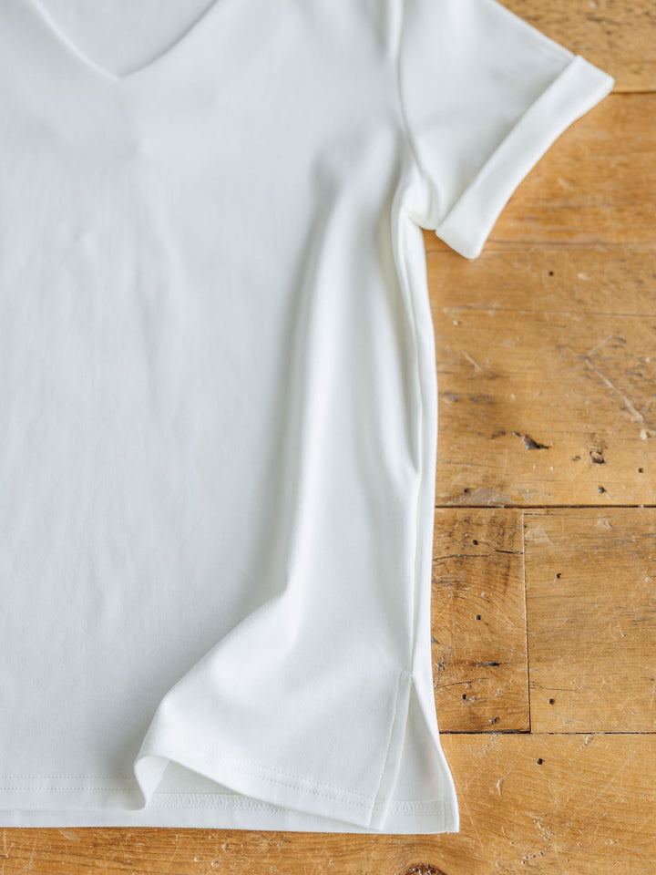 The Kinston by Savannah Sevens western life{&}style is an off-white short sleeve V-neck top with a slight hem fold and small side slit, displayed flat on a rustic wooden surface.