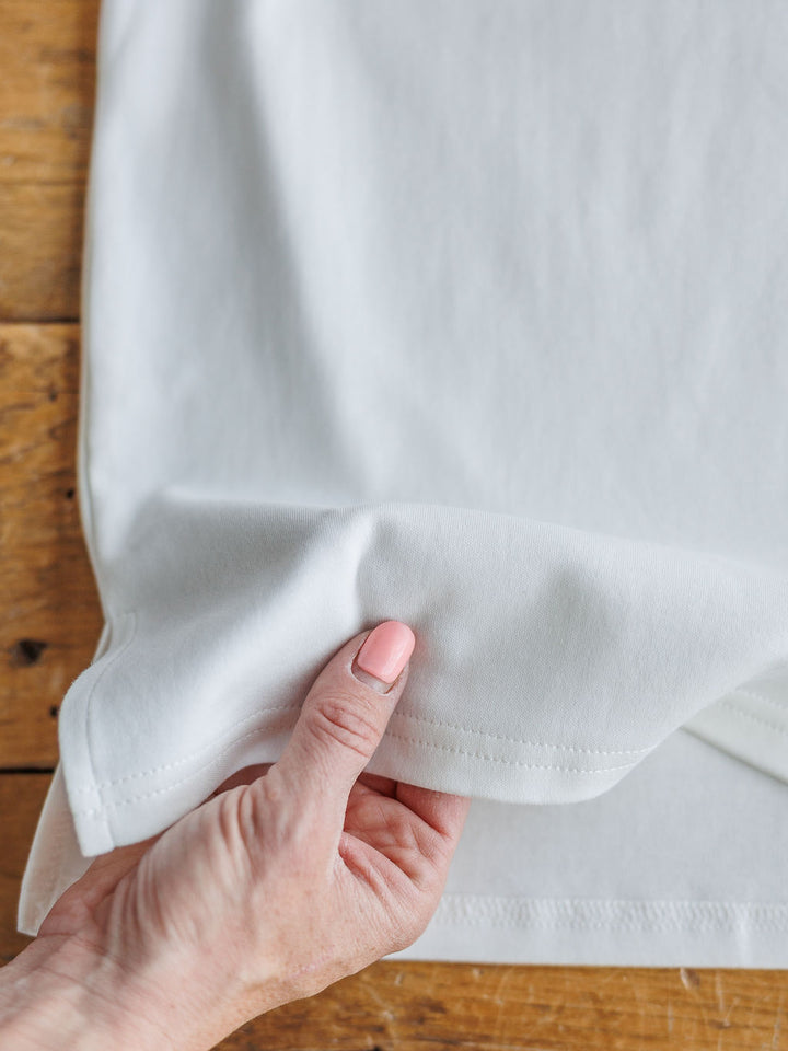 A hand with light pink nail polish lifts the edge of The Kinston off-white short sleeve top by Savannah Sevens western life{&}style, highlighting its thickness and texture on a wooden surface.