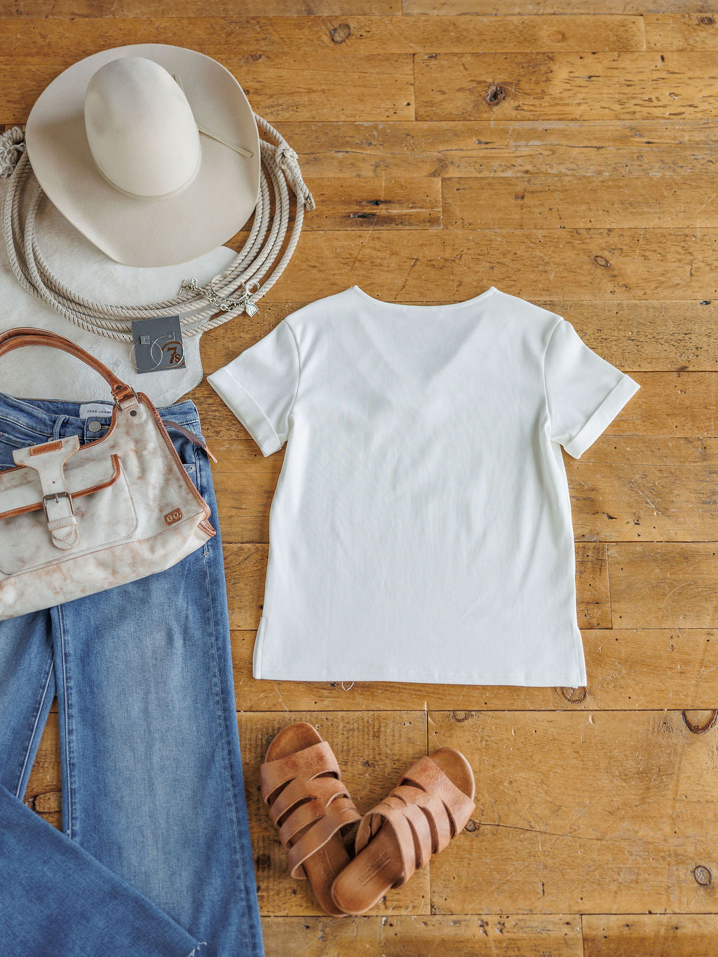A flat lay featuring The Kinston by Savannah Sevens western life{&}style: an off-white short sleeve top paired with blue jeans, tan sandals, a beige handbag, white cowboy hat, and a coiled rope on a wooden floor.