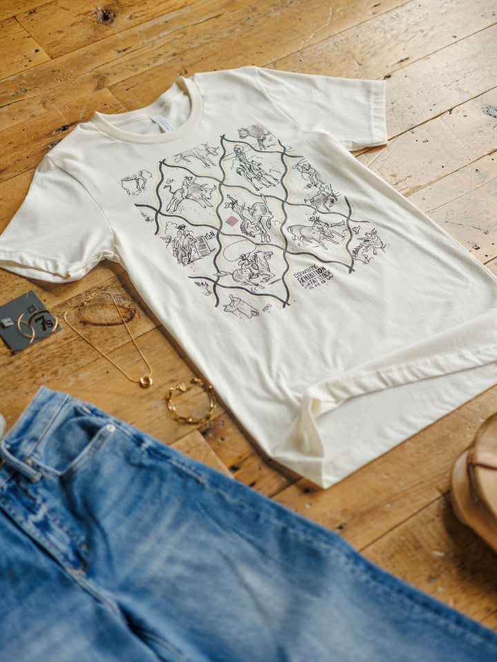 The Cowboy Exhibition white unisex cotton tee by Savannah Sevens western life{&}style is displayed on a wooden floor beside blue jeans, sandals, a gold bracelet and necklace. Detailed animal line art decorates the shirt.