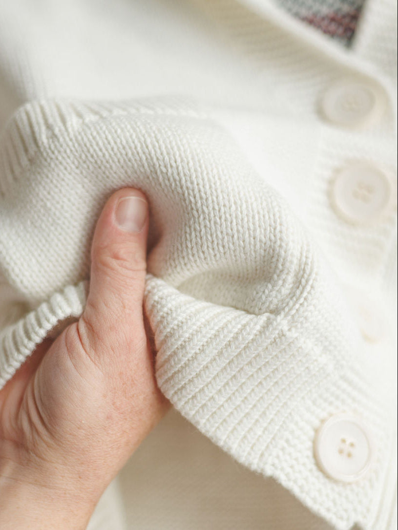A hand gently squeezes the sleeve of The American Classic {Exclusive} white knitted sweater by Savannah Sevens western life{&}style, showcasing its soft, textured fabric and large white buttons.