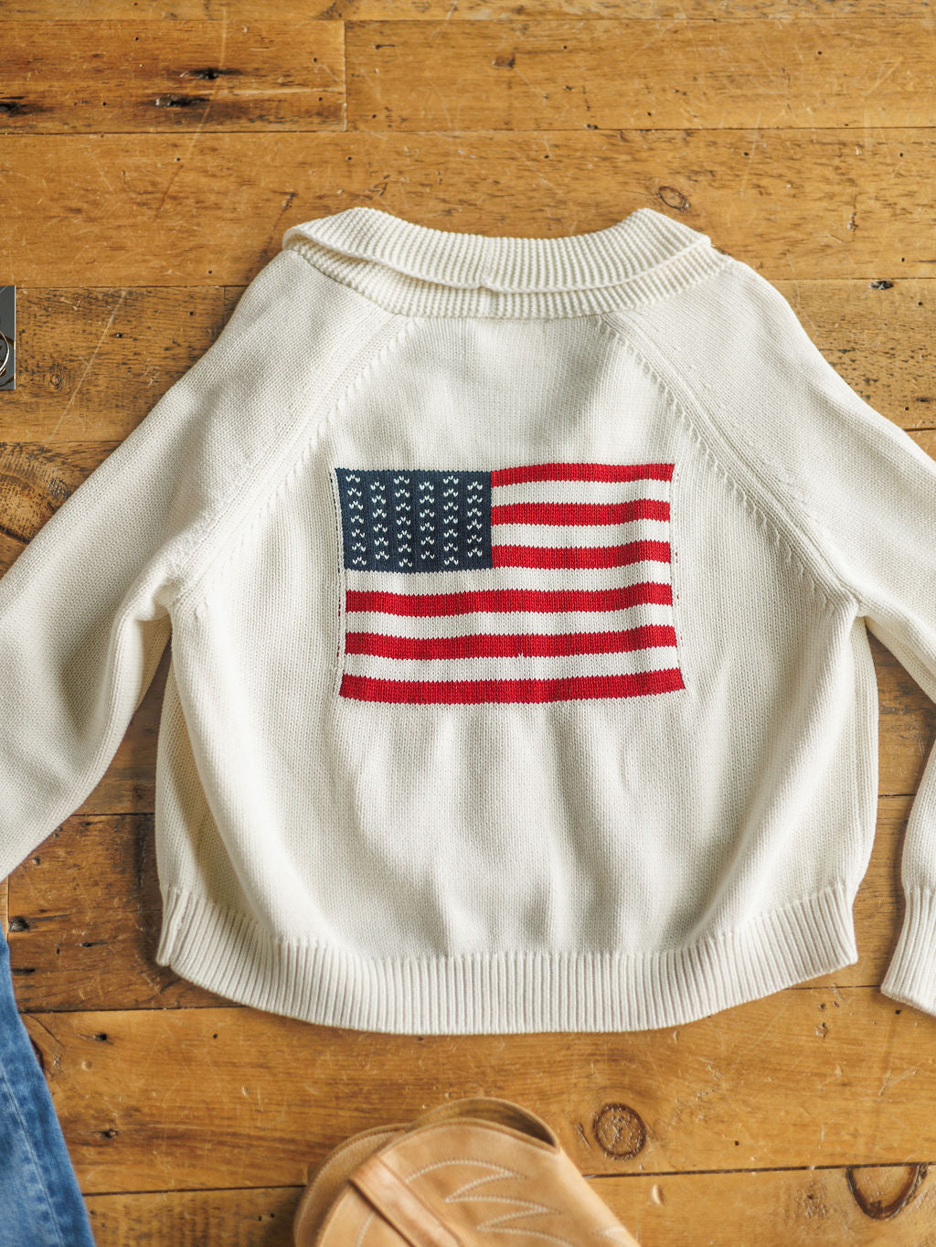 The American Classic {Exclusive} by Savannah Sevens western life{&}style is a white 100% cotton cardigan featuring an American flag design on the back, displayed on wood beside a sandal and blue denim—an exclusive Savannah Sevens piece.