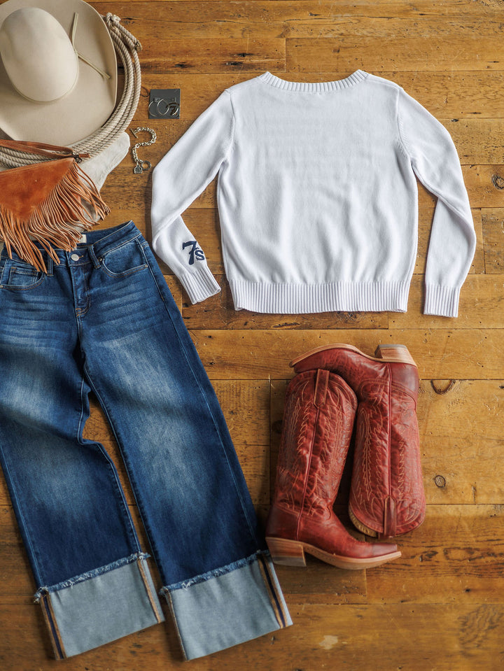 Flat lay of The American West {Exclusive} by Savannah Sevens western life{&}style: a white sweater, blue cuffed jeans, red cowboy boots, tan cowboy hat, tan fringe bag, and silver belt buckle on a wooden floor.