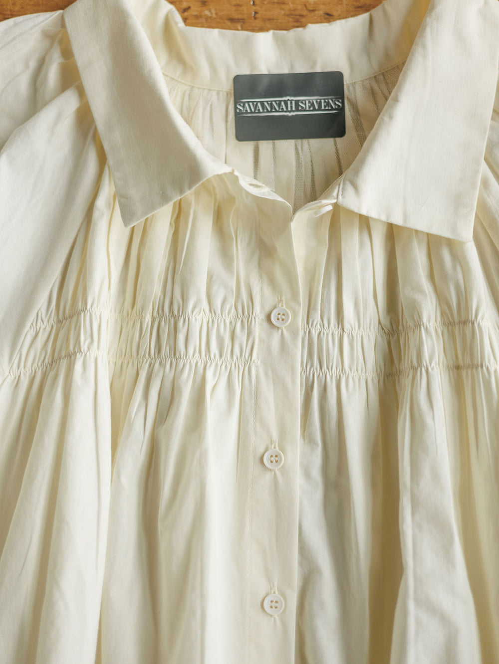 A close-up of The Annandale cream-colored cotton button-up blouse with gathered ruched detailing, collar, and subtle puff sleeves. Label: Savannah Sevens western life{&}style.