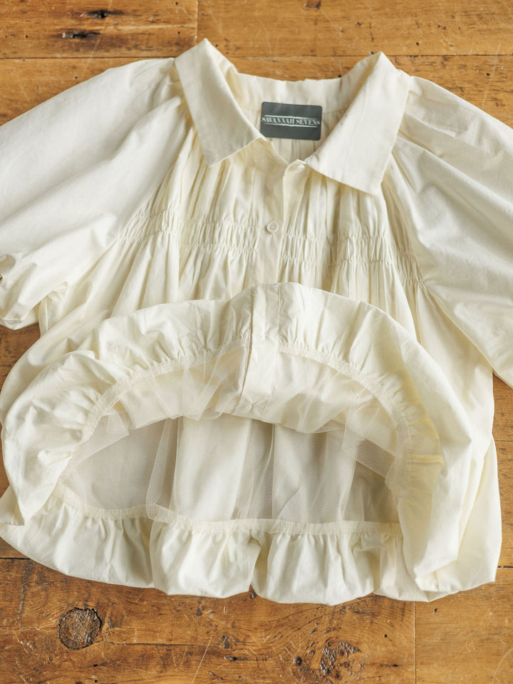 The Annandale by Savannah Sevens western life{&}style is a cream-colored cotton blouse with puff sleeves, gathered details, a ruffled hem, collared neckline, and button-down front, displayed on a wooden surface.