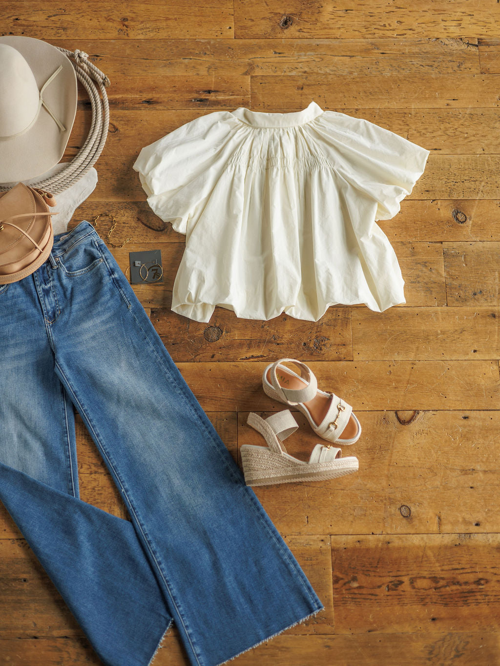 Flat lay on wood: The Annandale wide-leg jeans by Savannah Sevens western life{&}style, white puff sleeve blouse, beige wedge sandals, wide-brim hat, tan crossbody bag, and stud earrings.