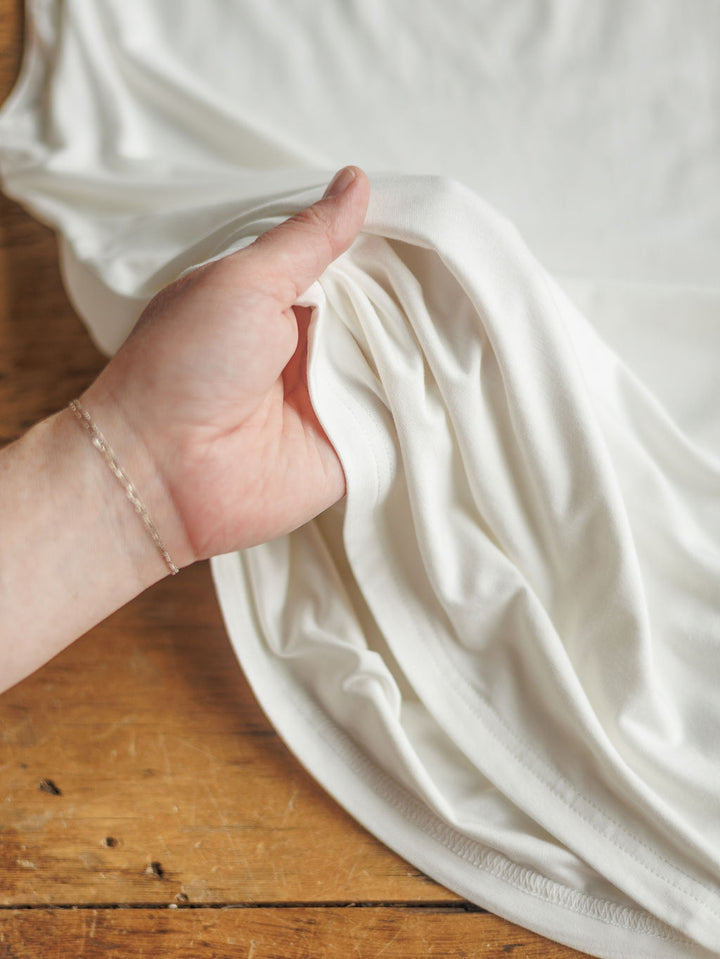 A hand gently lifts The Renton from Savannah Sevens western life{&}style, showcasing its buttery soft white fabric and smooth drape above a wooden surface.
