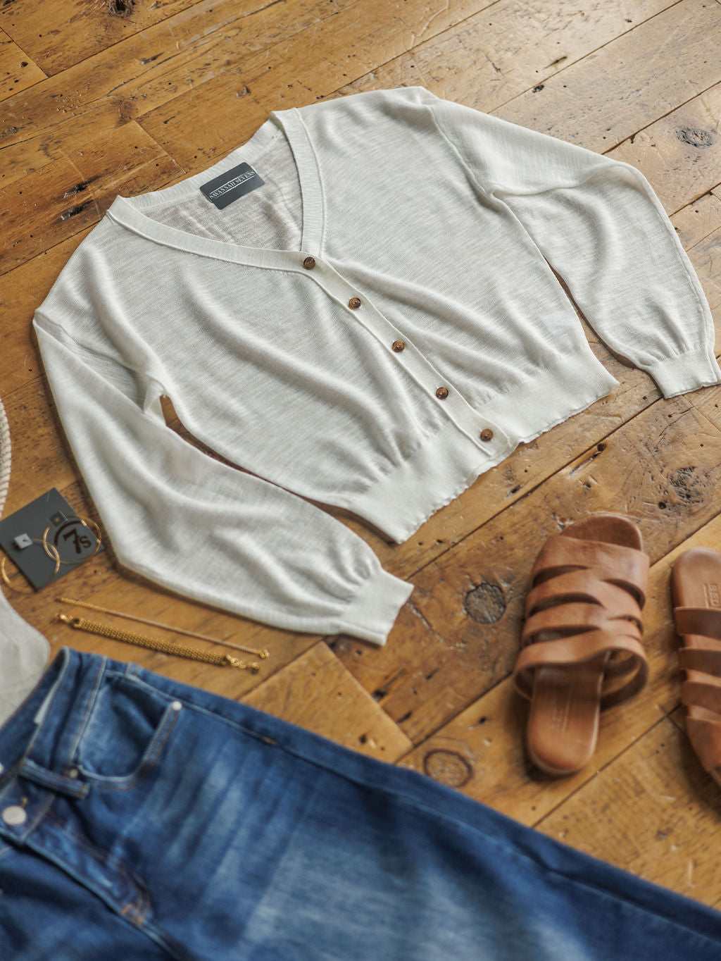 The Caney sweater by Savannah Sevens western life{&}style, in light beige woven knit with long sleeves, is displayed on a wooden floor beside blue jeans, brown sandals, a gold bracelet, and keys.