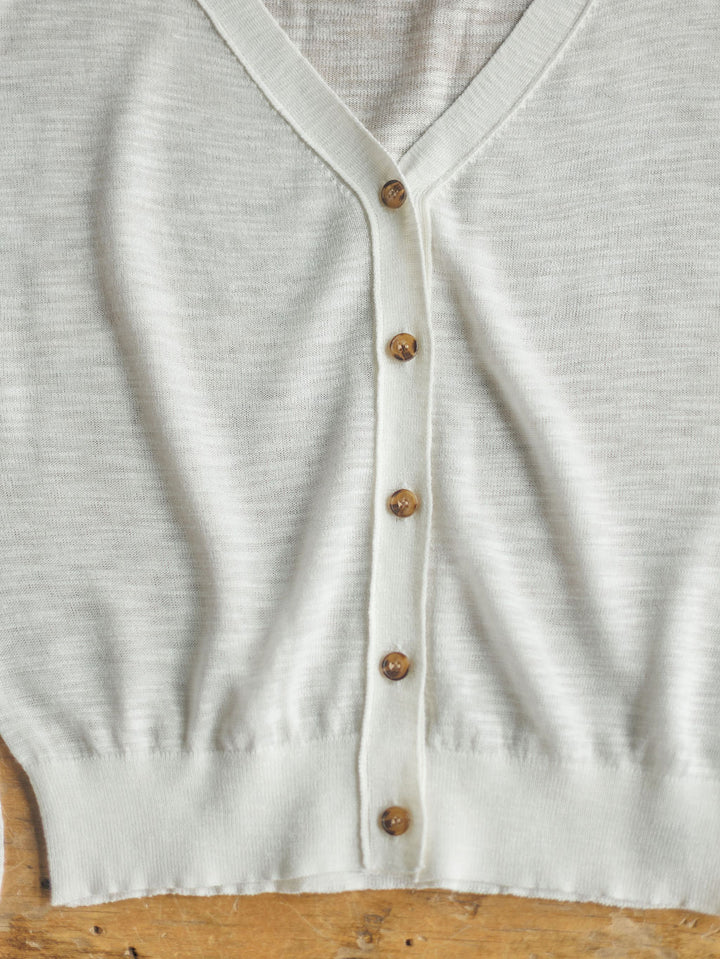 A close-up of The Caney by Savannah Sevens western life{&}style, a sheer white long sleeve V-neck knit cardigan with six brown buttons, laid flat on a wooden surface.