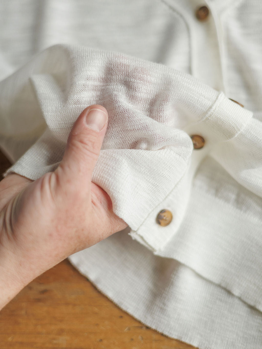A hand pinches the semi-sheer white fabric of The Caney long sleeve button up by Savannah Sevens western life{&}style, showing its lightweight, slightly translucent texture and brown button details against a wooden background.