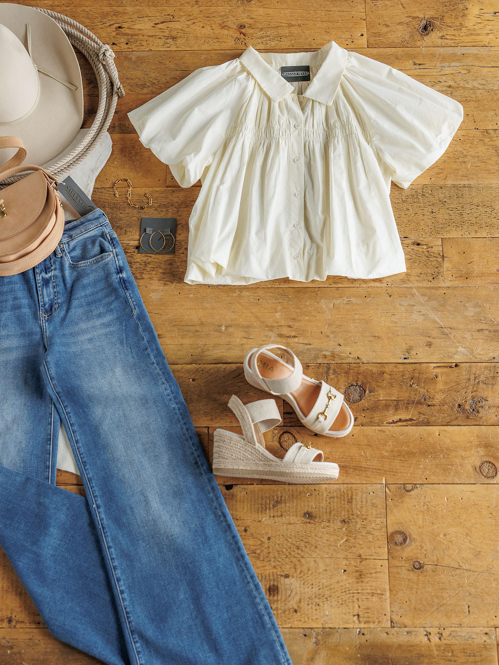 A flat lay of a casual outfit on a wooden floor featuring The Annandale blouse by Savannah Sevens western life{&}style, blue jeans, white wedge sandals, a tan hat, tan crossbody bag, and hoop earrings.