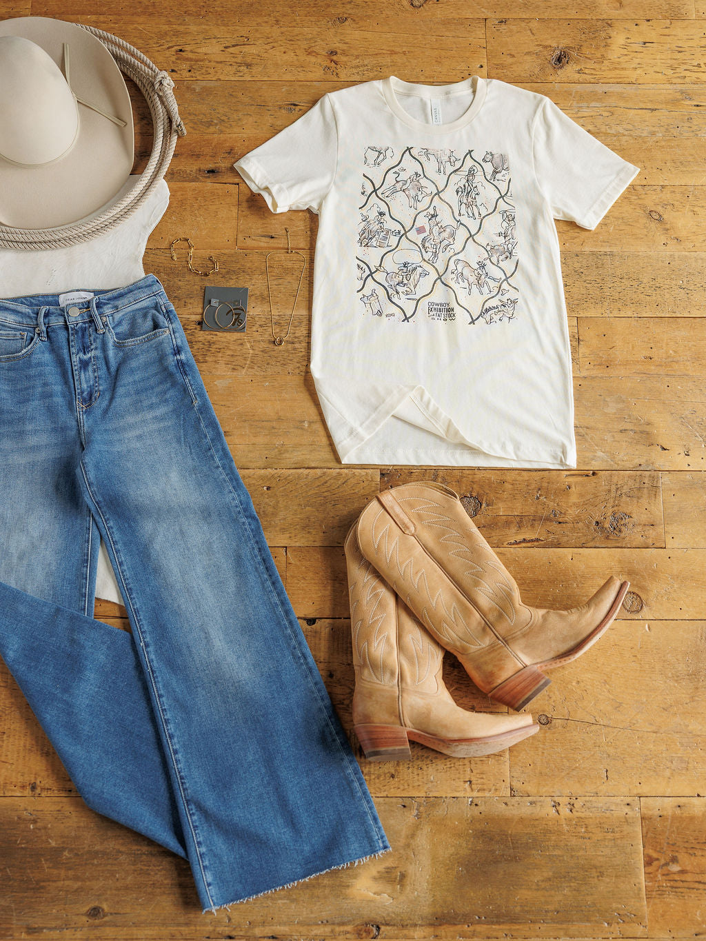 Savannah Sevens western life{&}style's "The Cowboy Exhibition" features blue jeans, a floral western tee, tan cowboy boots, wide-brim hat, belt, and silver jewelry including hoops and necklace—laid out on a wooden floor.