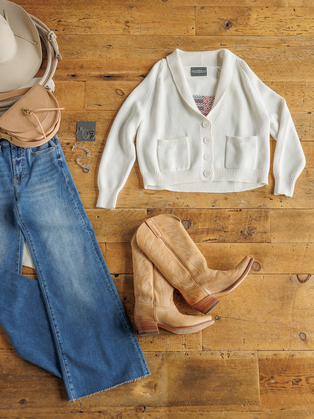 Flat lay of Savannah Sevens western life{&}style's exclusive: The American Classic cardigan, blue jeans, tan cowboy boots, beige wide-brim hat, tan crossbody purse, silver necklace and bracelet on a wooden floor.