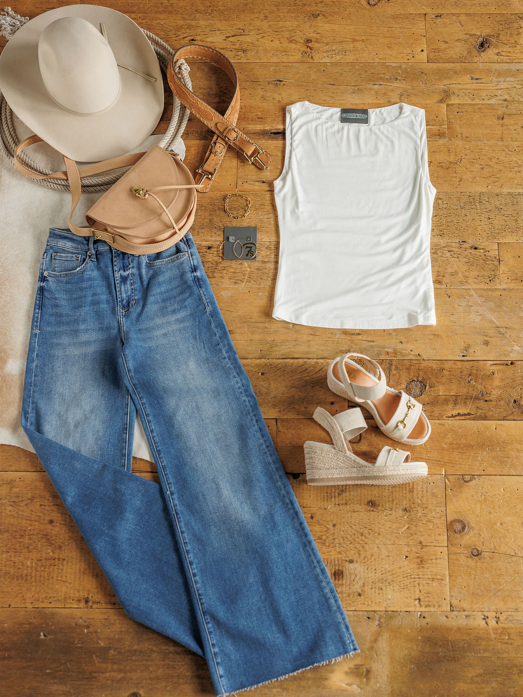 Flat lay of The Renton by Savannah Sevens western life&style: wide-brimmed hat, tan crossbody bag, light brown belt, buttery soft white modal tank, blue wide-leg jeans, beige wedge sandals, and earrings on a wooden floor.