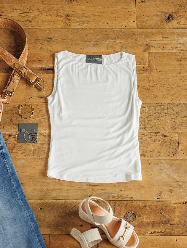 The Renton by Savannah Sevens western life{&}style—a buttery soft white modal tank—is laid flat on a wooden floor, styled with a brown belt, gold bracelet, denim jeans, small camera, and white heeled sandals.