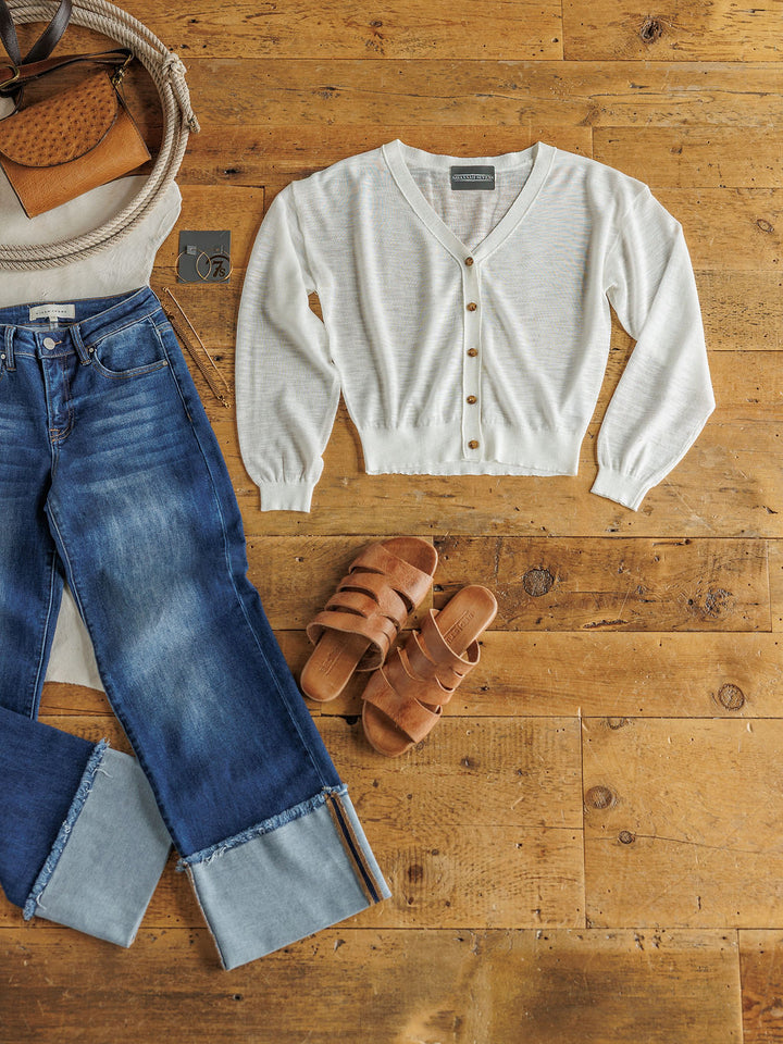 Flat lay of The Caney sheer white long sleeve button up cardigan by Savannah Sevens western life{&}style, blue wide cuff denim jeans, brown slide sandals, and a tan woven handbag with leather accent on a wooden floor.