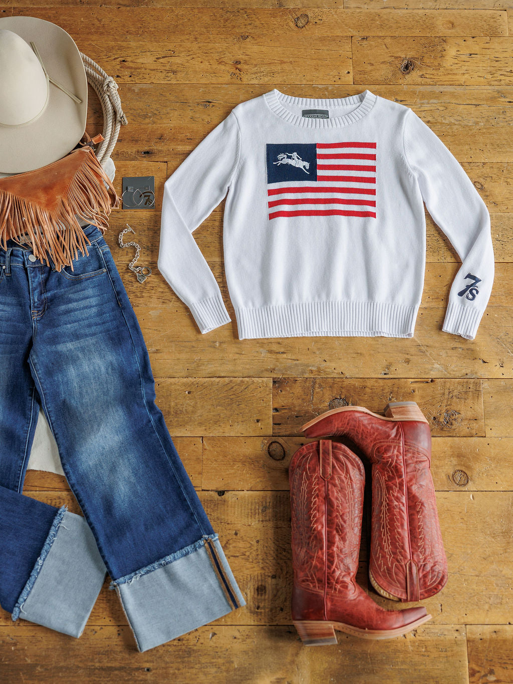 Flat lay featuring The American West {Exclusive} by Savannah Sevens western life{&}style: American flag knit top with horse design, cuffed blue jeans, red cowboy boots, white hat, fringed bag, and silver jewelry on a wooden floor.