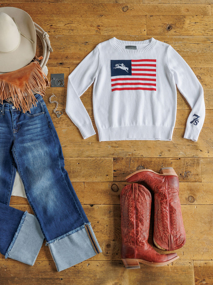 Flat lay featuring The American West {Exclusive} by Savannah Sevens western life{&}style: American flag knit top with horse design, cuffed blue jeans, red cowboy boots, white hat, fringed bag, and silver jewelry on a wooden floor.