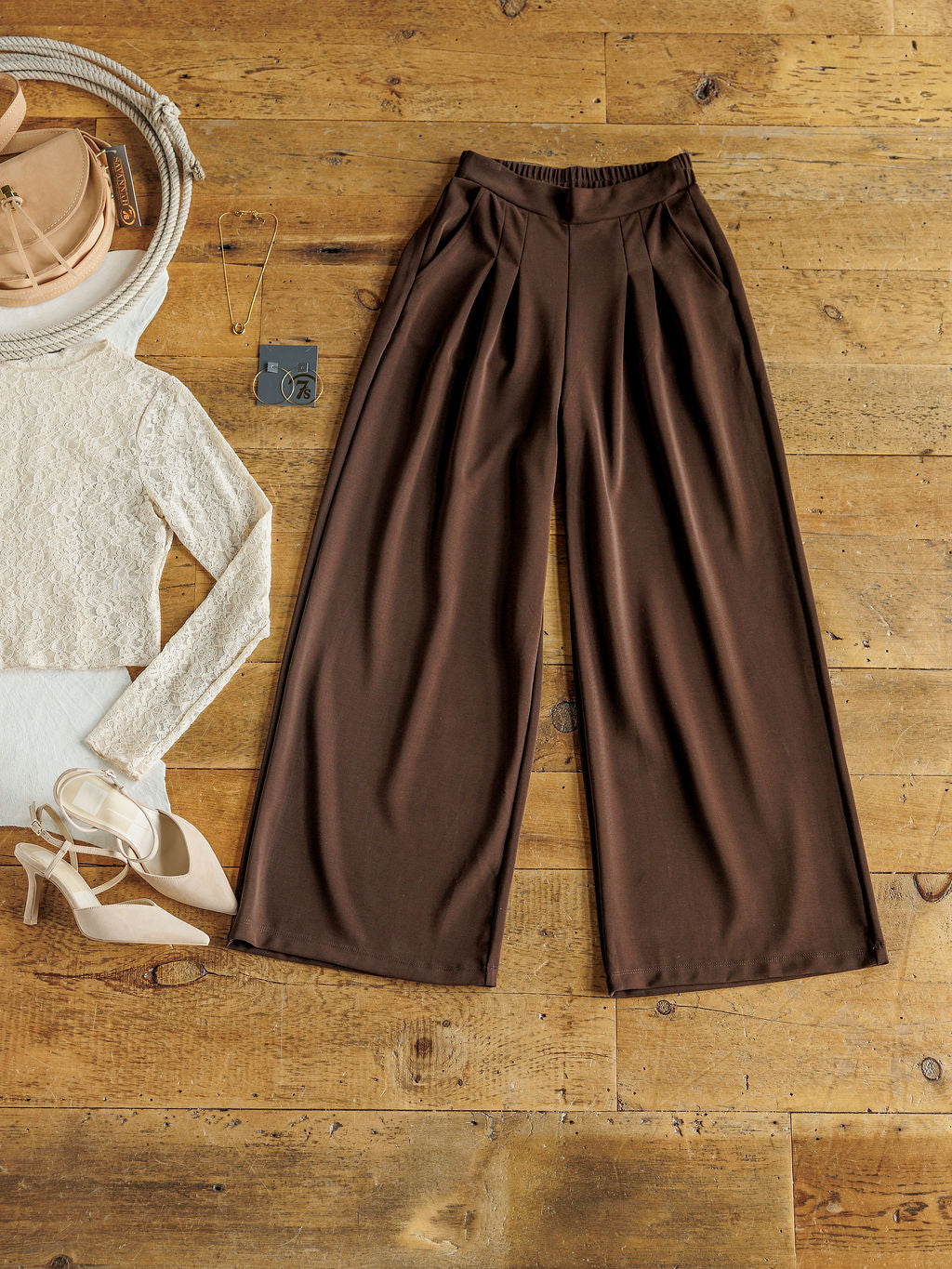 The Kline wide leg pants by Savannah Sevens western life{&}style are styled on a wooden floor with a cream knit top, beige heels, pink purse, sunglasses, necklace, and keychain.
