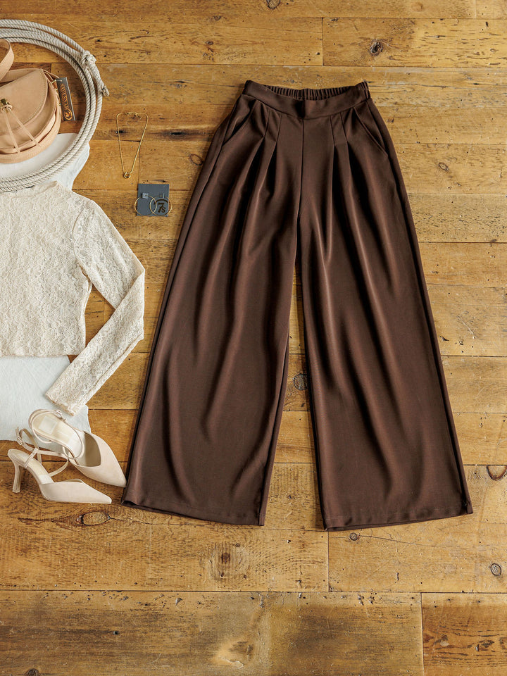 The Kline wide leg pants by Savannah Sevens western life{&}style are styled on a wooden floor with a cream knit top, beige heels, pink purse, sunglasses, necklace, and keychain.