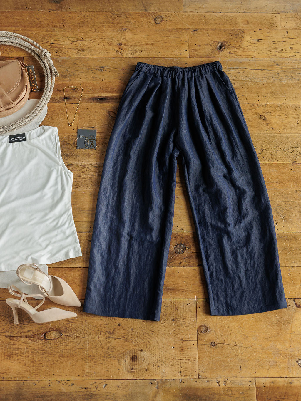 The Meridan high waisted wide leg dark denim pants by Savannah Sevens western life{&}style, laid flat with a white sleeveless top, tan handbag, nude heels, gold necklace, and gray cardholder on a wooden floor.