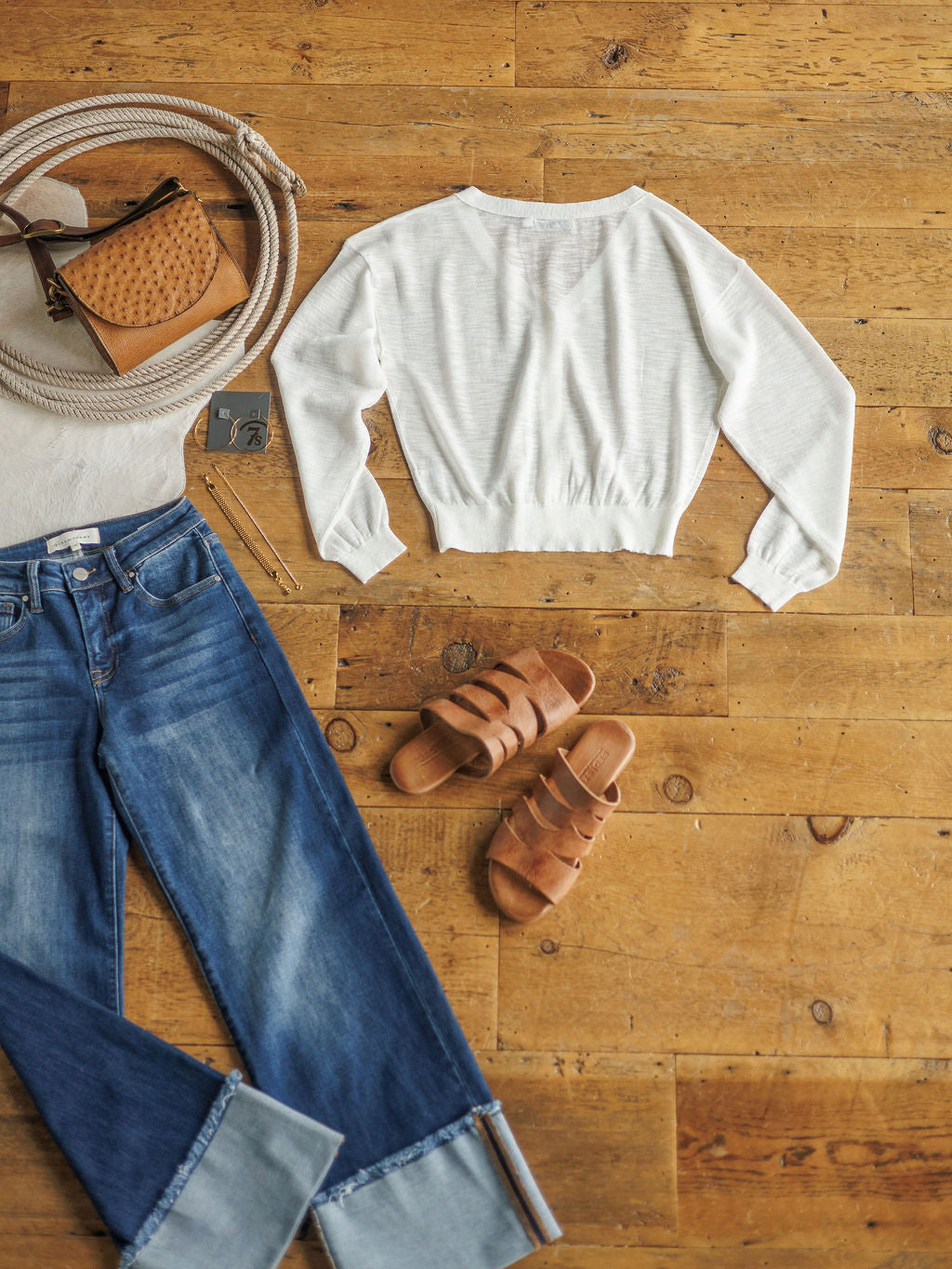 A casual outfit from Savannah Sevens western life{&}style includes The Caney blue cuffed jeans, a sheer white cardigan, brown slide sandals, tan crossbody bag, coiled belt, silver necklace, and sunglasses on a wooden floor.