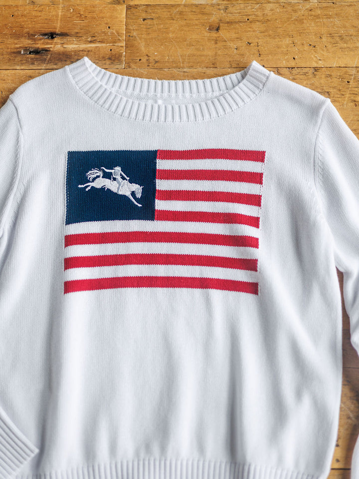 A white "The American West {Exclusive}" knit sweater by Savannah Sevens western life{&}style features a blue field with a bucking bronco horse silhouette embroidered on the front. The long sleeve top is displayed flat on a wooden surface.