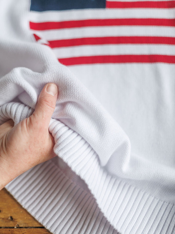 A hand gently grasps the ribbed edge of The American West {Exclusive} embroidered long sleeve top in a white American flag knit with red stripes and blue, by Savannah Sevens western life{&}style, on a wooden surface.