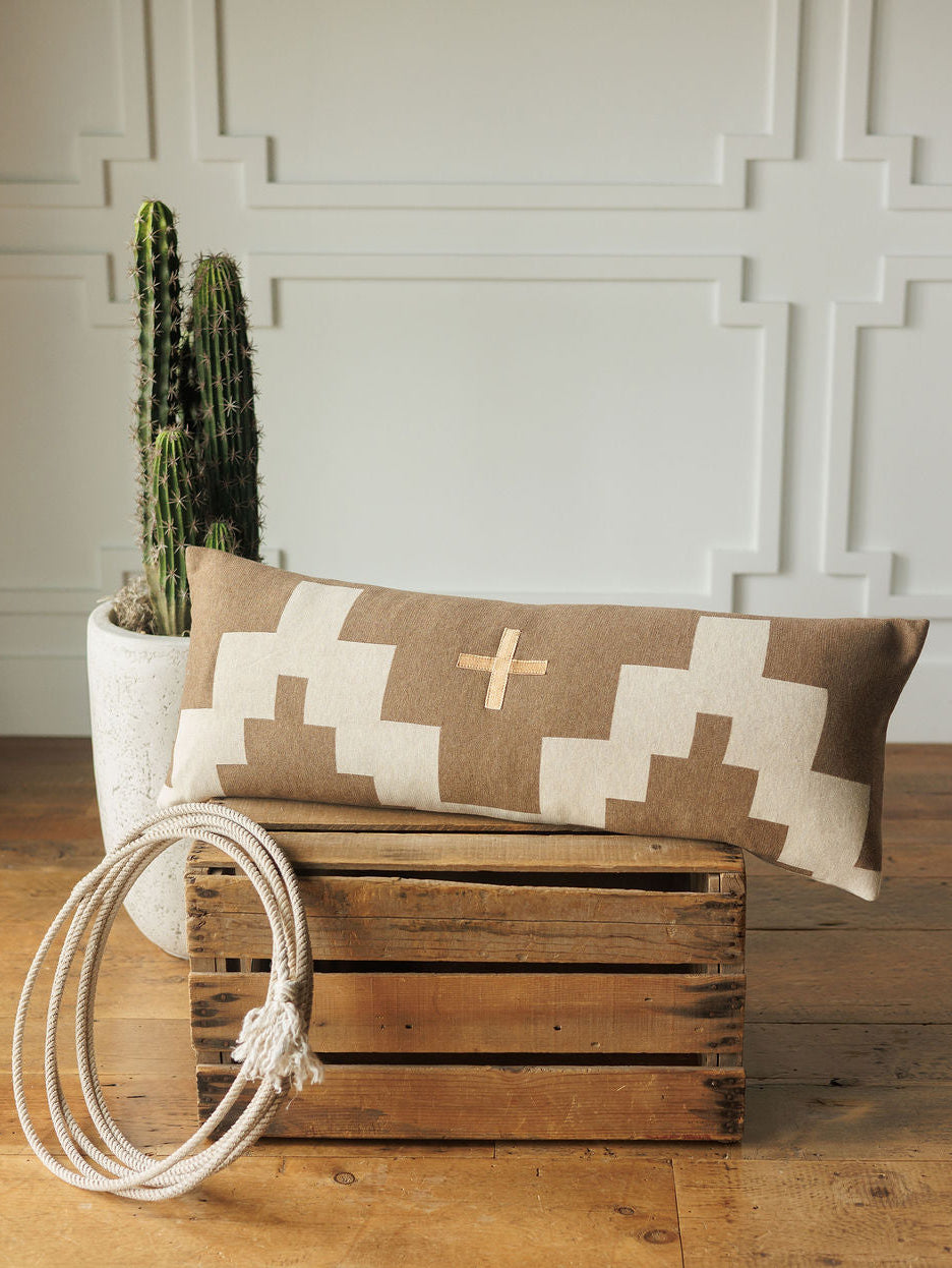 The Presidio Lumbar Pillow Cover {Tan} by Savannah Sevens western life{&}style, with a geometric cross pattern, rests on a wooden crate beside coiled white rope and a cactus in a white planter, set against a paneled wall.