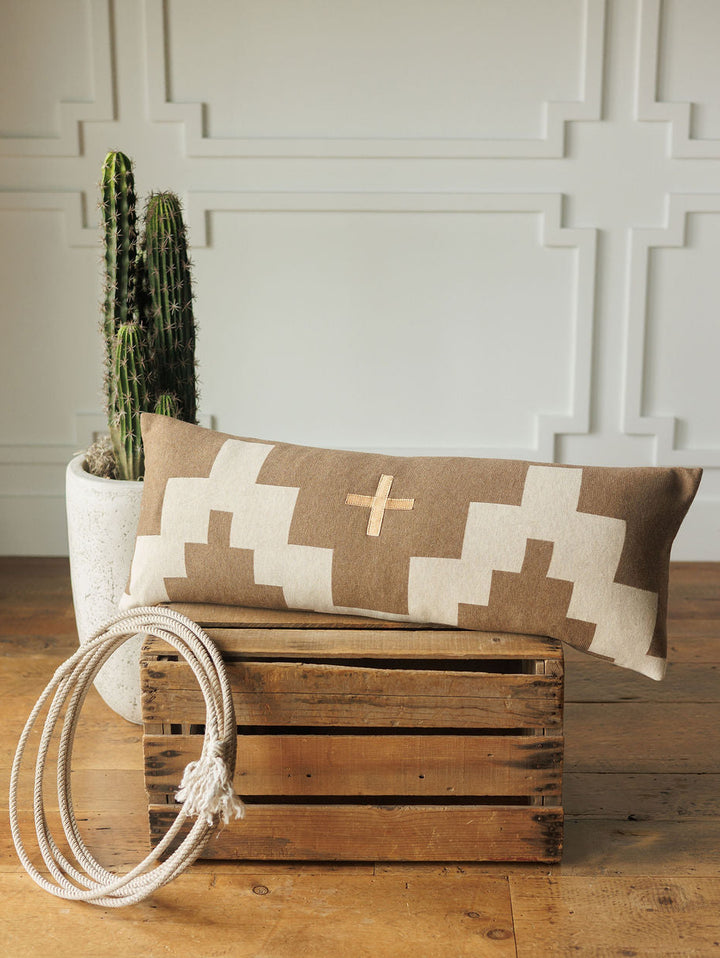 The Presidio Lumbar Pillow Cover {Tan} by Savannah Sevens western life{&}style, with a geometric cross pattern, rests on a wooden crate beside coiled white rope and a cactus in a white planter, set against a paneled wall.