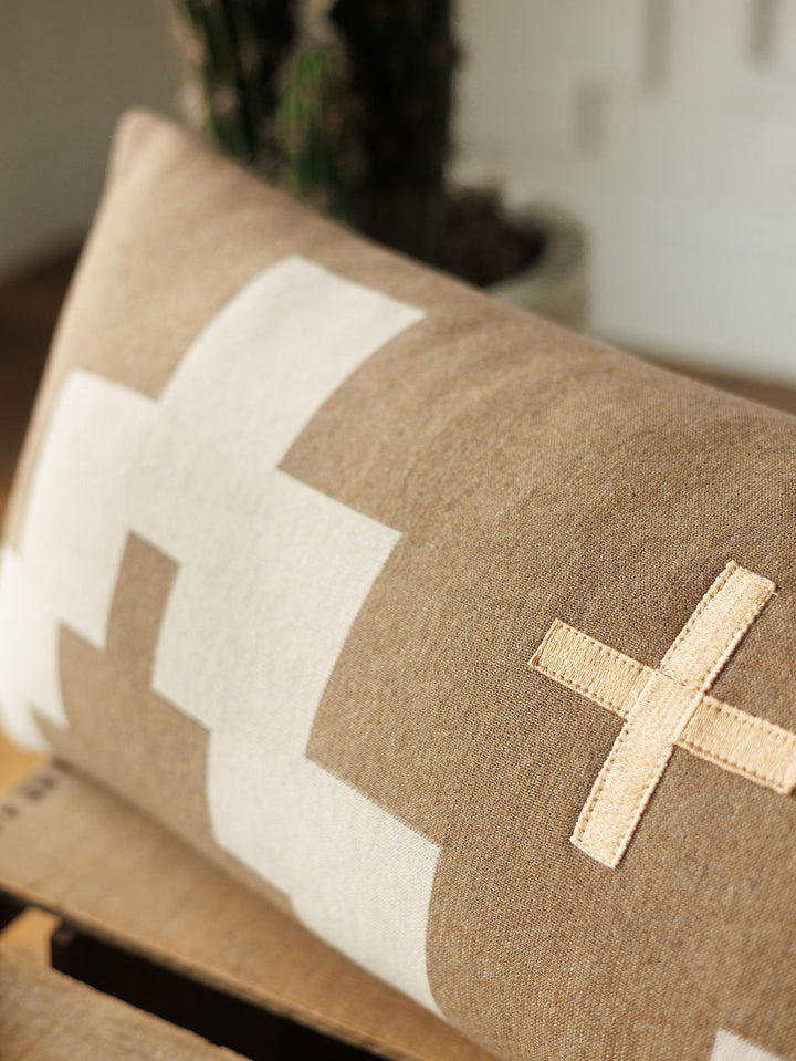 The Presidio Lumbar Pillow Cover {Tan} by Savannah Sevens western life{&}style features bold white geometric cross patterns and a beige stitched cross, displayed on a wooden surface with a blurred background.