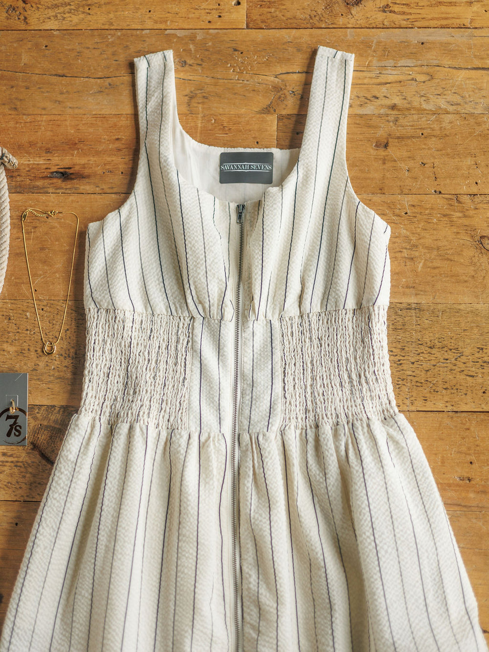 The Palomar by Savannah Sevens western life{&}style, a sleeveless cotton dress with thin blue stripes and a smocked waist, is shown laid flat on wood with a gold necklace and clothing tag beside it.