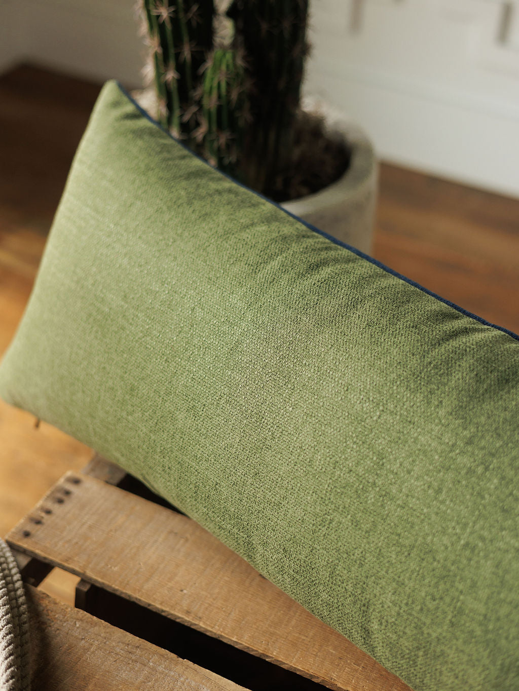 The Presidio Lumbar Pillow Cover in Midnight Navy from Savannah Sevens western life{&}style rests on a wooden surface, with a potted cactus in the background and sunlight illuminating the scene.
