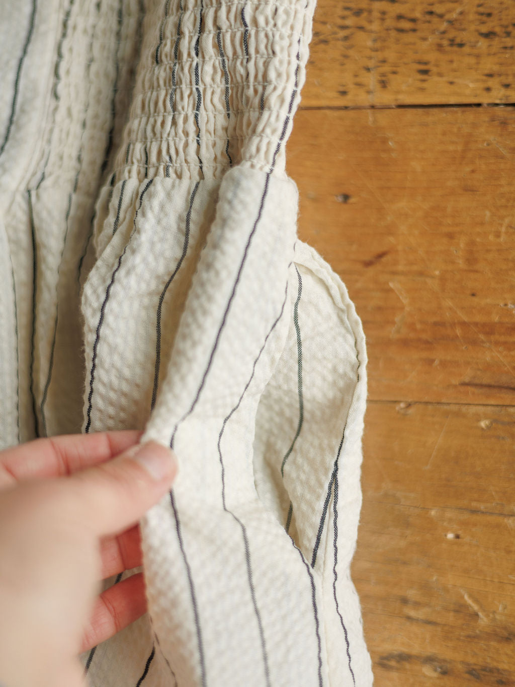 A hand holds the pocket of The Palomar, a cream-colored midi dress with vertical black stripes by Savannah Sevens western life{&}style, displayed on a wooden surface.