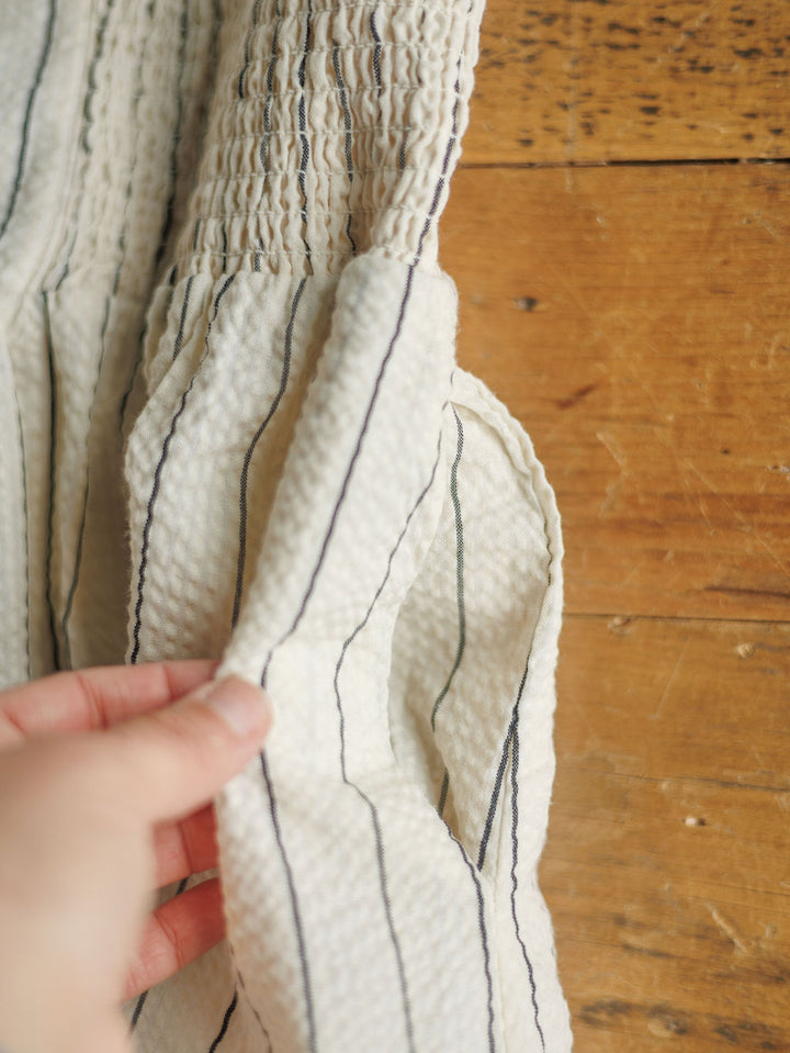 A hand holds the pocket of The Palomar, a cream-colored midi dress with vertical black stripes by Savannah Sevens western life{&}style, displayed on a wooden surface.