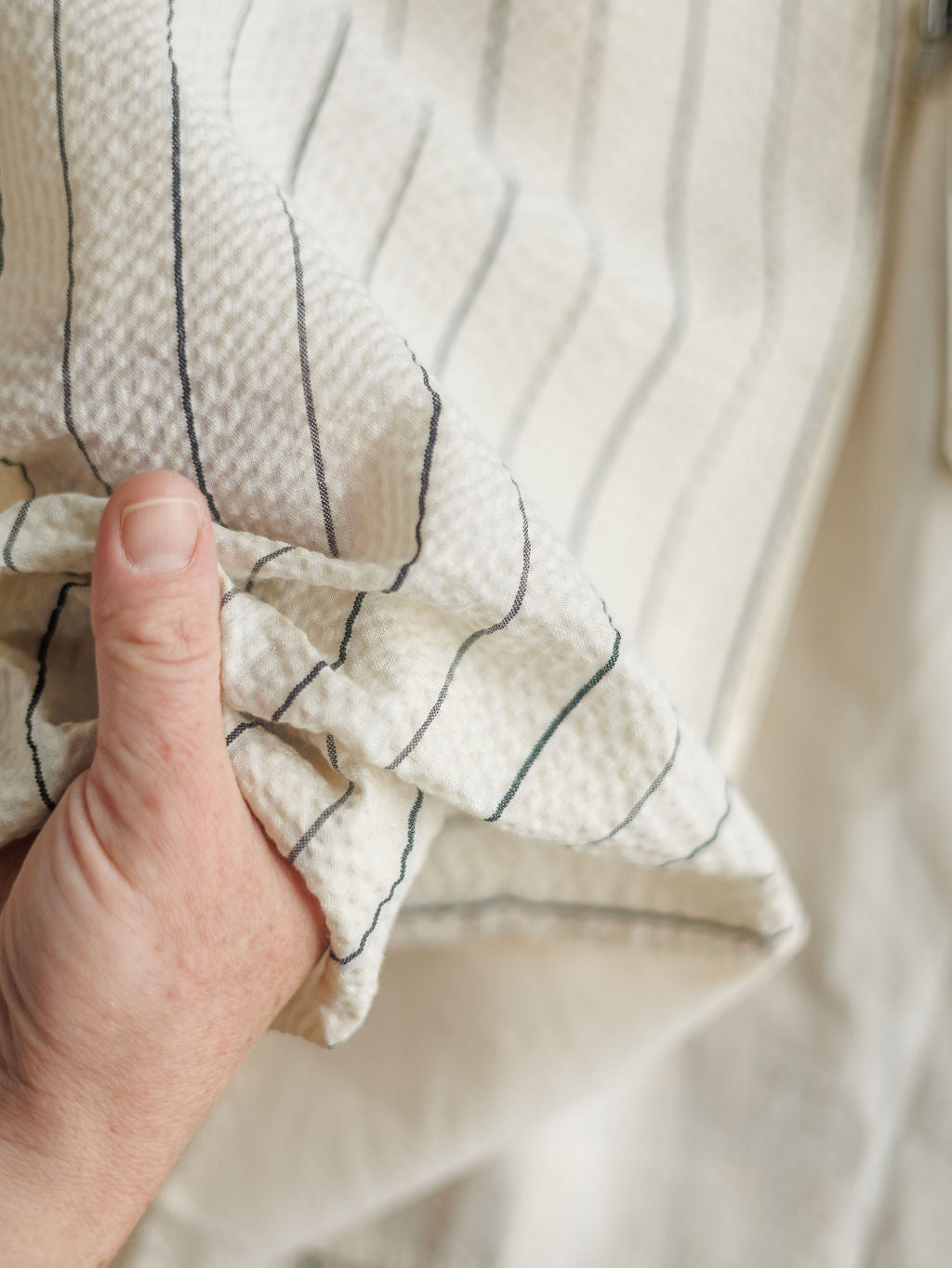 A hand gently holds The Palomar, a textured white midi dress with thin dark stripes from Savannah Sevens western life{&}style, against a soft, light background.