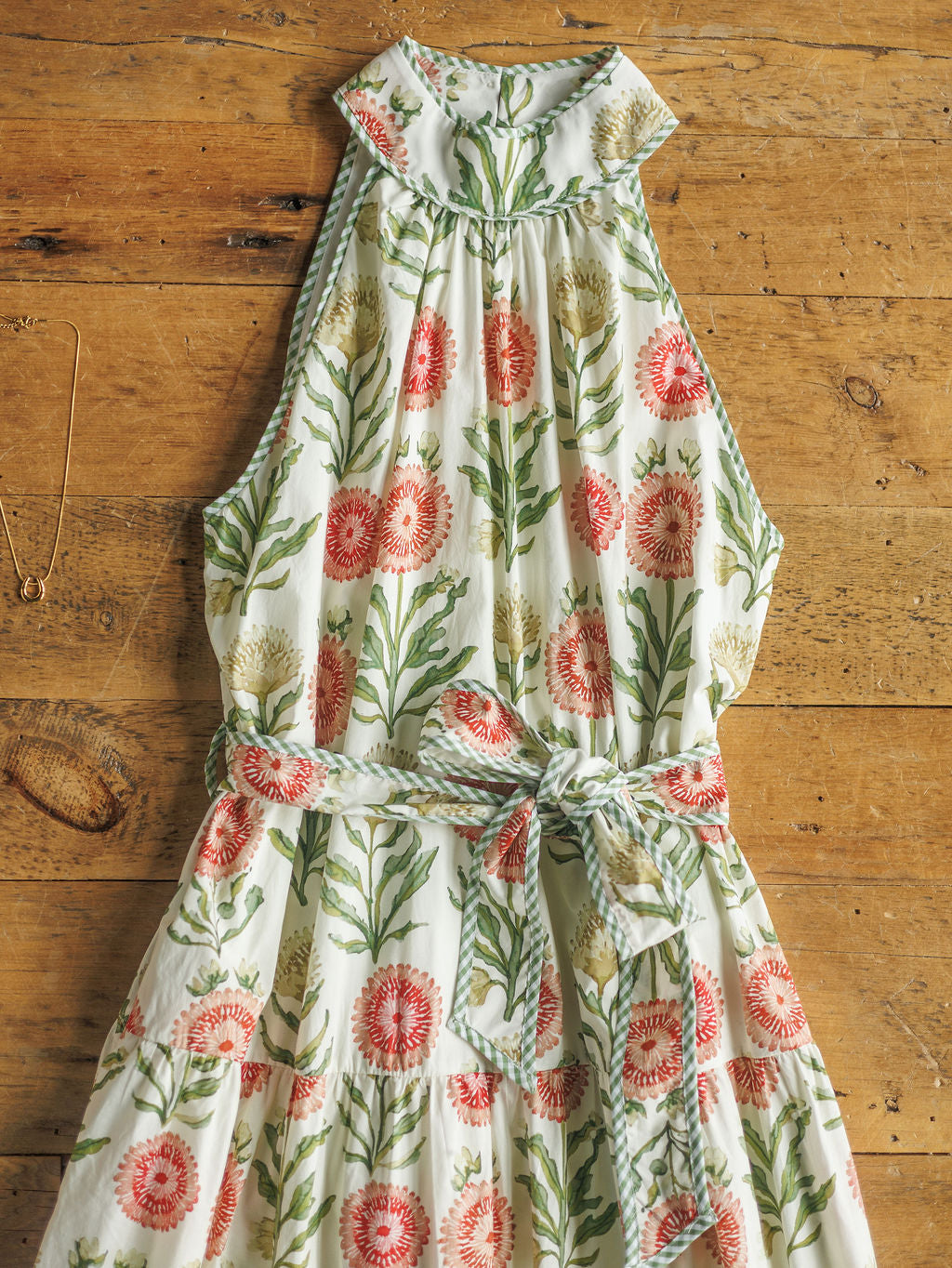 The Marianna dress by Savannah Sevens western life{&}style is a cotton, full-length floral piece with a high neckline and waist tie, showcasing pink and white flowers with green leaves on a rustic wooden background.