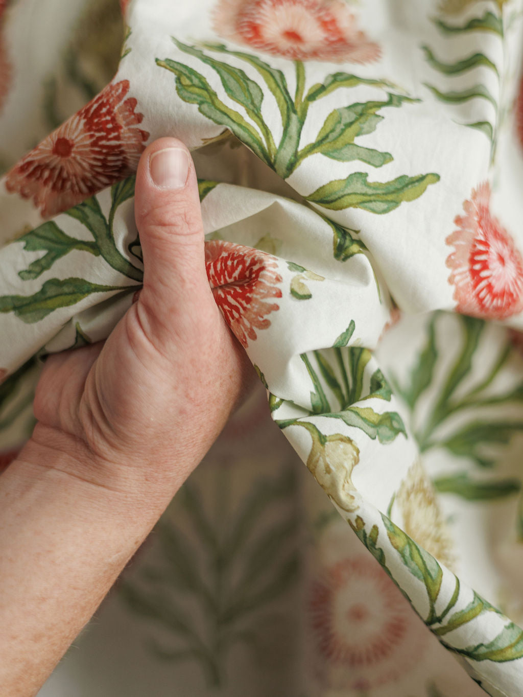 A hand holds the soft cotton fabric of The Marianna by Savannah Sevens western life{&}style, featuring green leaves and red-orange floral patterns that showcase its vibrant full-length dress design.