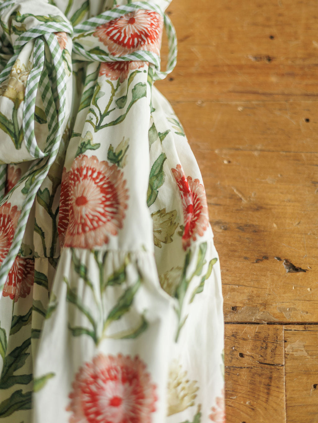 A close-up of The Marianna, a full-length cotton dress by Savannah Sevens western life{&}style, featuring a red and green floral pattern and gathered at the waist with a green and white striped sash on rustic wood.
