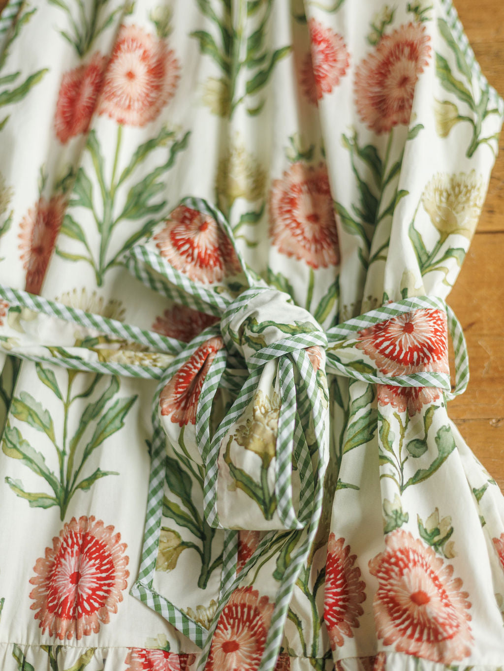A close-up of The Marianna dress by Savannah Sevens western life{&}style in red, green, and cream with floral and leaf prints, a halter neck, a tied waist belt, and green striped trim.