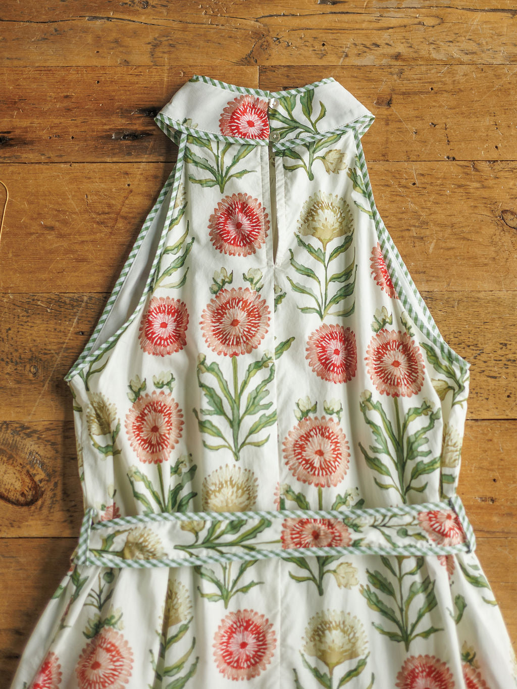 The Marianna by Savannah Sevens western life{&}style is a sleeveless halter dress with a high neckline, pink and white floral print with green leaves, and green-and-white striped edging, laid flat on a rustic wooden surface.