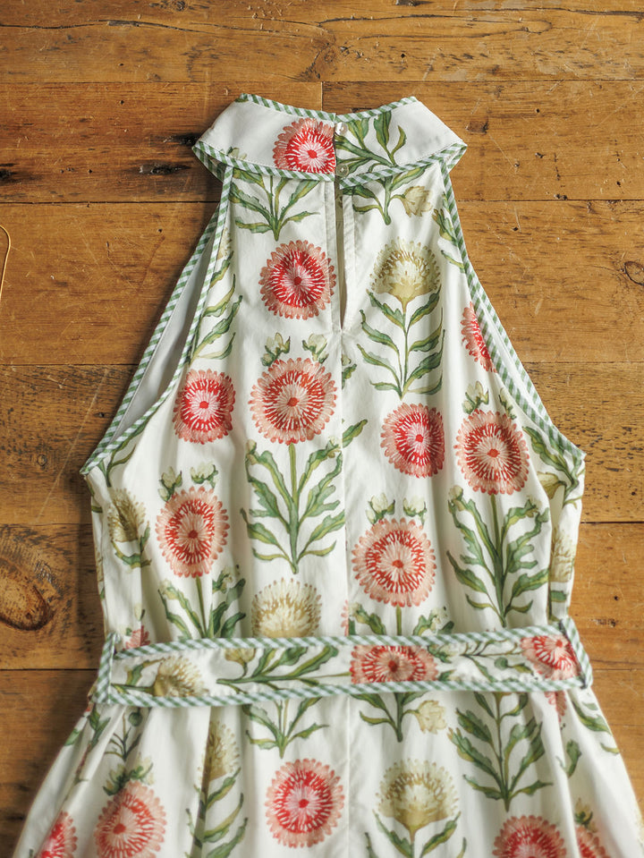 The Marianna by Savannah Sevens western life{&}style is a sleeveless halter dress with a high neckline, pink and white floral print with green leaves, and green-and-white striped edging, laid flat on a rustic wooden surface.