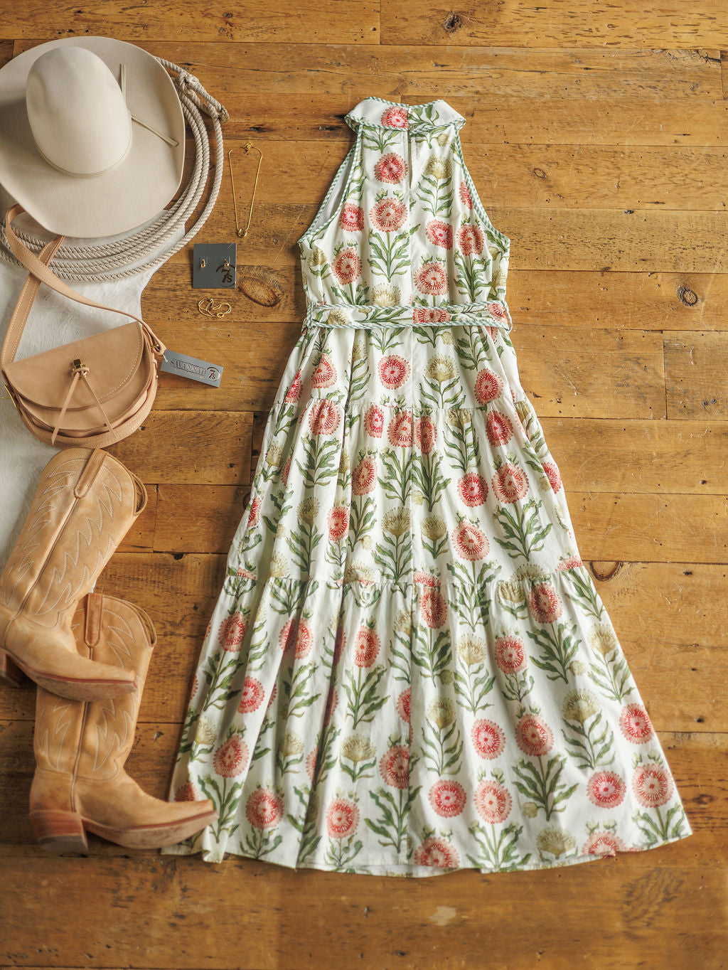 The Marianna by Savannah Sevens western life{&}style is styled on a wooden floor with tan cowboy boots, a beige hat, a tan crossbody purse, a white sweater, and gold jewelry arranged beside the sleeveless floral halter neck dress.