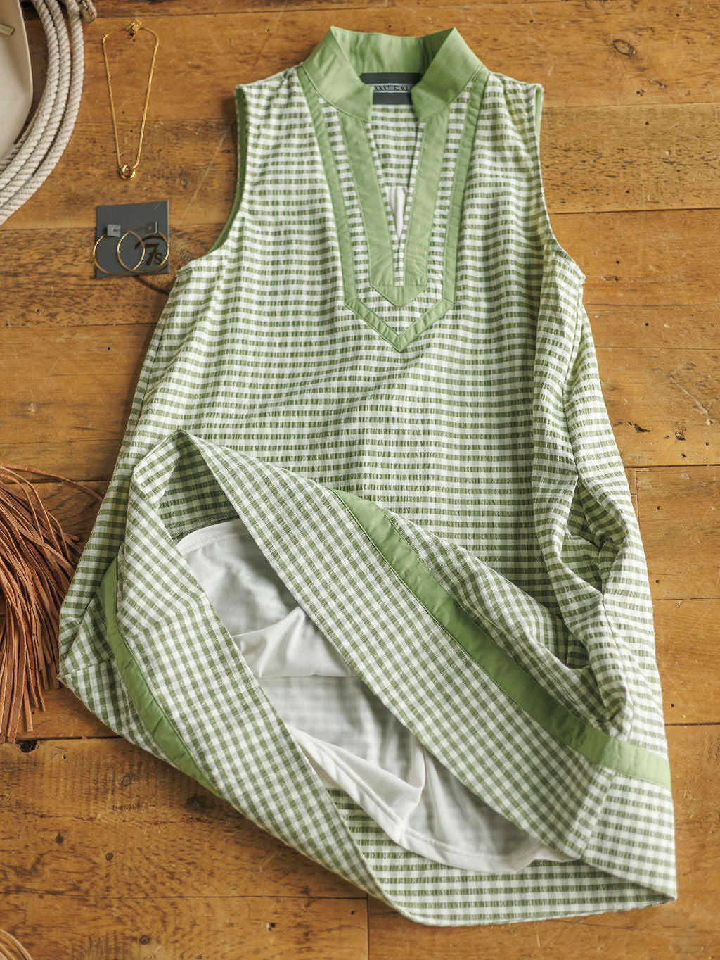 The Halstad by Savannah Sevens western life{&}style, a sleeveless green and white gingham V-neck dress with green trim, is styled on a wood surface beside a necklace, earrings, hat, and fringed brown bag for a modern Western vibe.