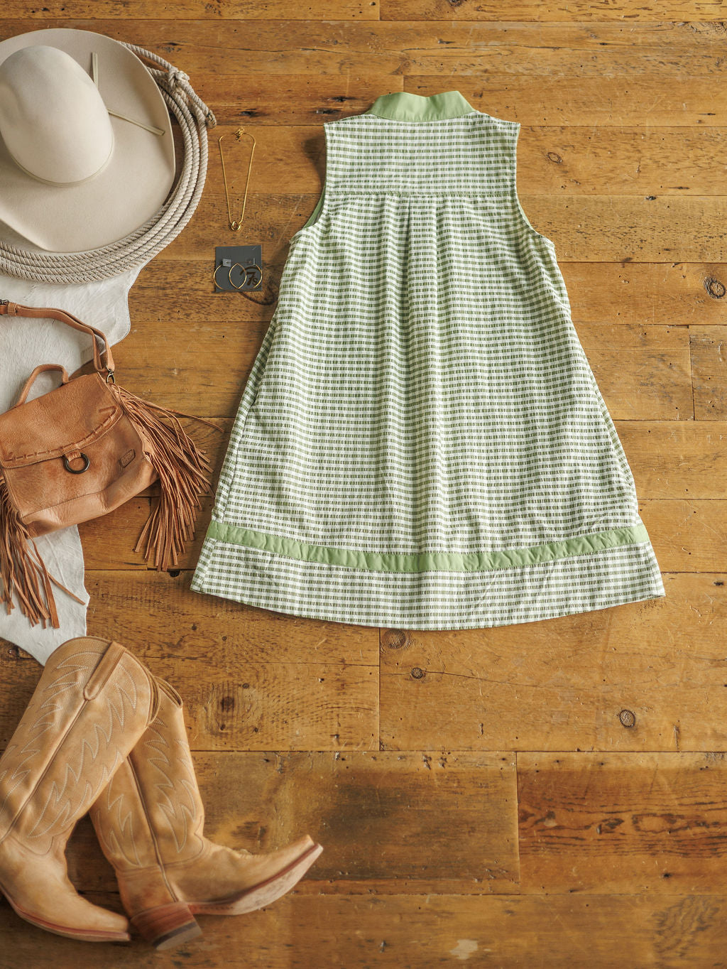 The Halstad by Savannah Sevens western life{&}style, a sleeveless green and white gingham dress, is styled on a wooden floor with a white cowboy hat, tan fringed bag, jewelry, scarf, and tan cowboy boots.