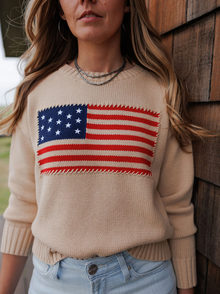 A woman wears The Original {Exclusive} by Savannah Sevens western life{&}style—an American flag 100% cotton sweater—with light blue jeans, standing against a wooden wall. Only her torso and part of her face are visible.