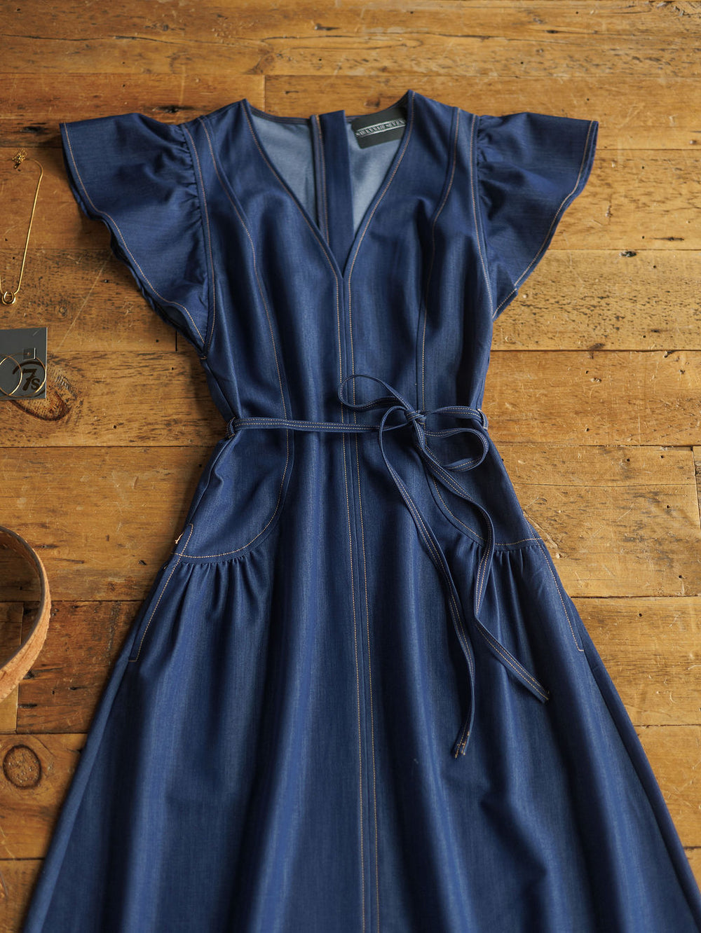 The Navassa by Savannah Sevens western life{&}style is a blue denim midi dress with short ruffled sleeves, V-neck, front seam details, and a petite waist tie, laid flat on a wooden surface.