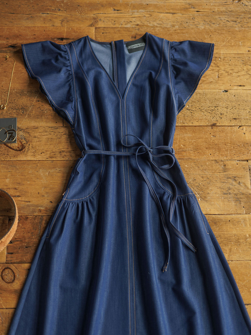 The Navassa by Savannah Sevens western life{&}style is a blue denim midi dress with short ruffled sleeves, V-neck, front seam details, and a petite waist tie, laid flat on a wooden surface.